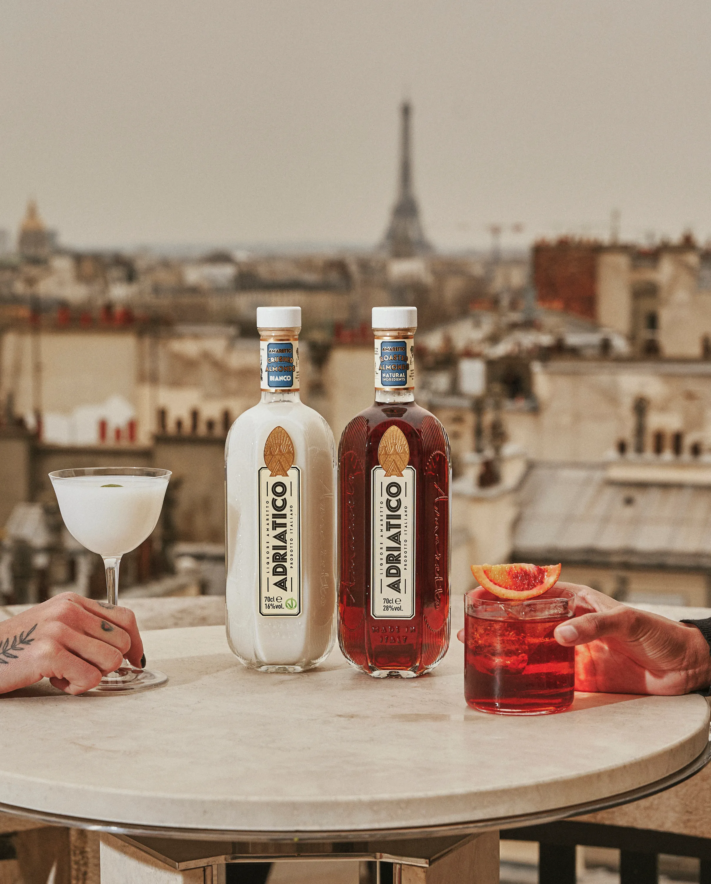 Two bottles of Adriatico liqueur on a round table with a white cocktail on the left and a red cocktail with an orange slice on the right, cityscape background with the Eiffel Tower.