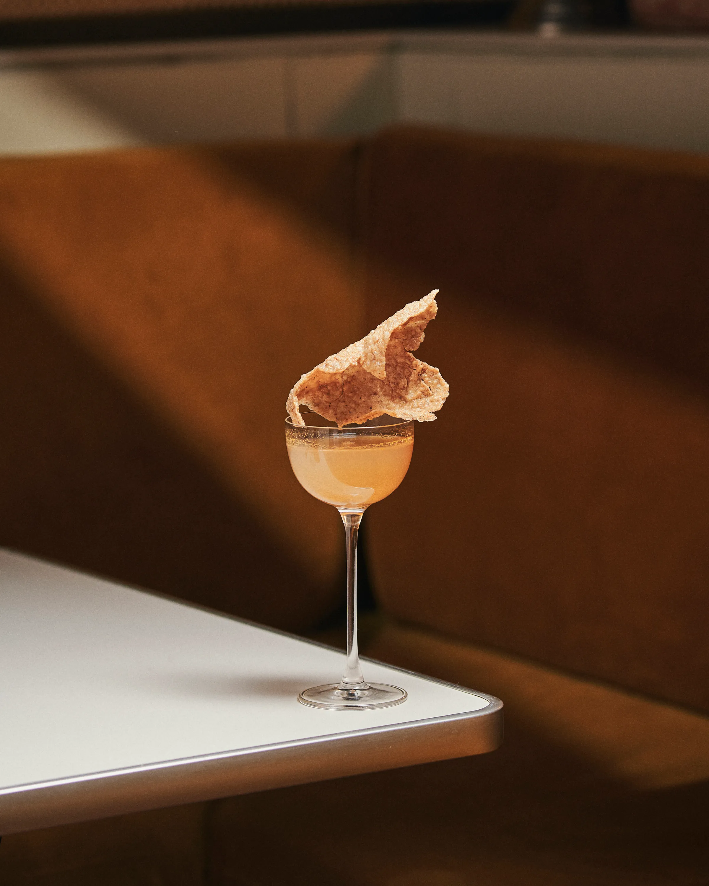 Cocktail in a coupe glass with a large crispy garnish on top, placed on a white table with brown cushioned seating in the background.