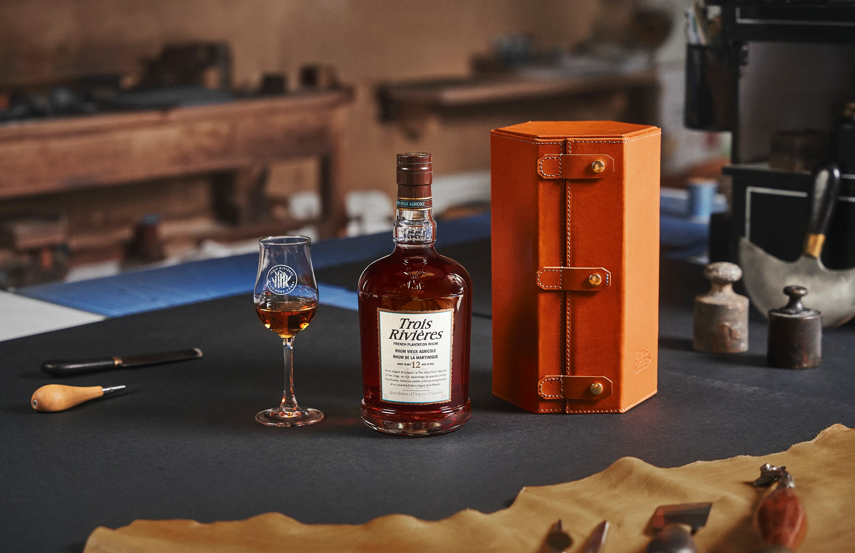 Bottle of Trois Rivières French plantation rum with a filled glass and an orange leather case on a dark table in a workshop setting.