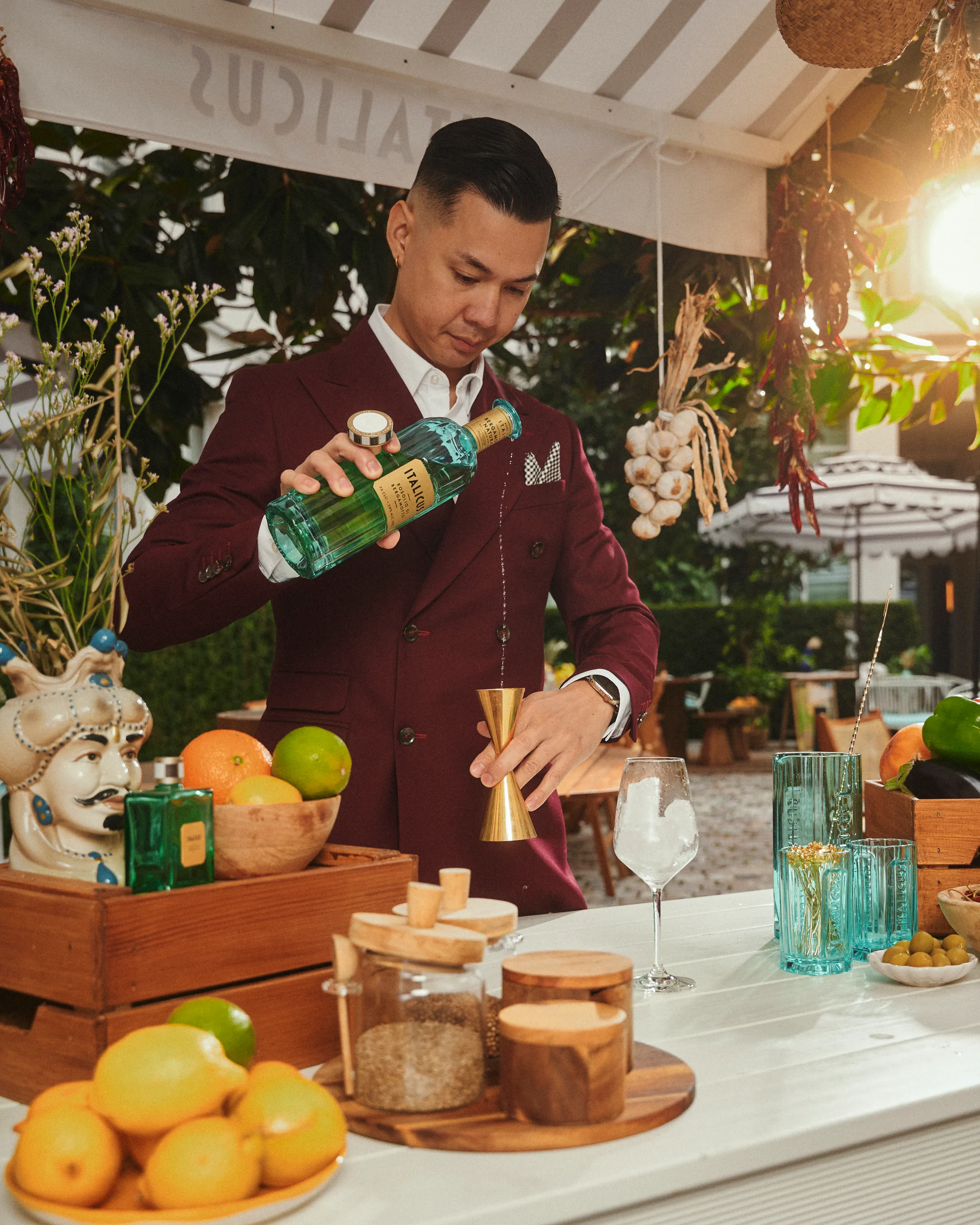 Man in burgundy suit pouring Italicus liqueur into a gold jigger at an outdoor bar with fruits, jars, and glassware on the counter.