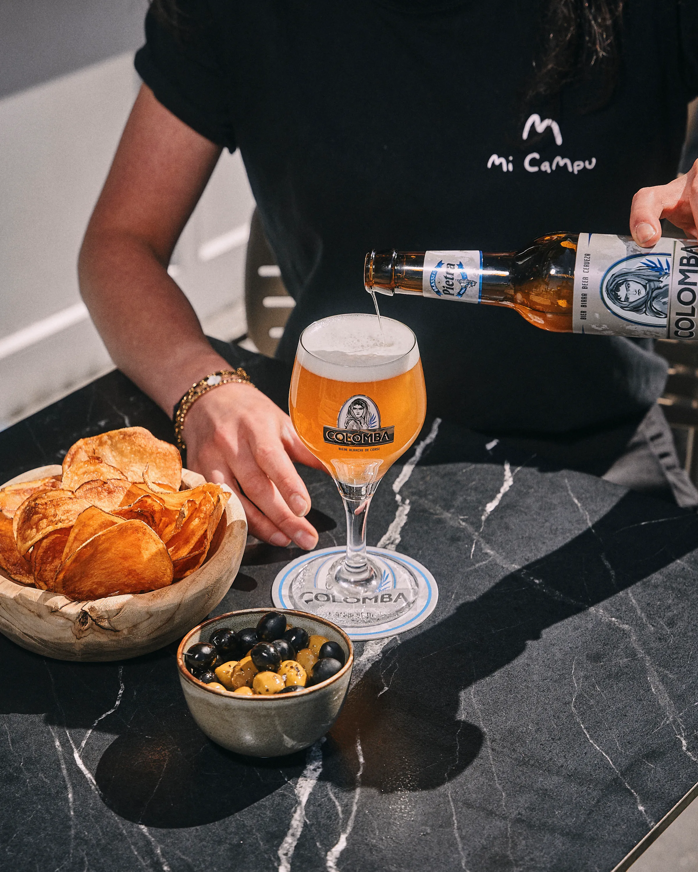 Person pouring Colomba beer into a branded glass on a black marble table next to bowls of potato chips and mixed olives.