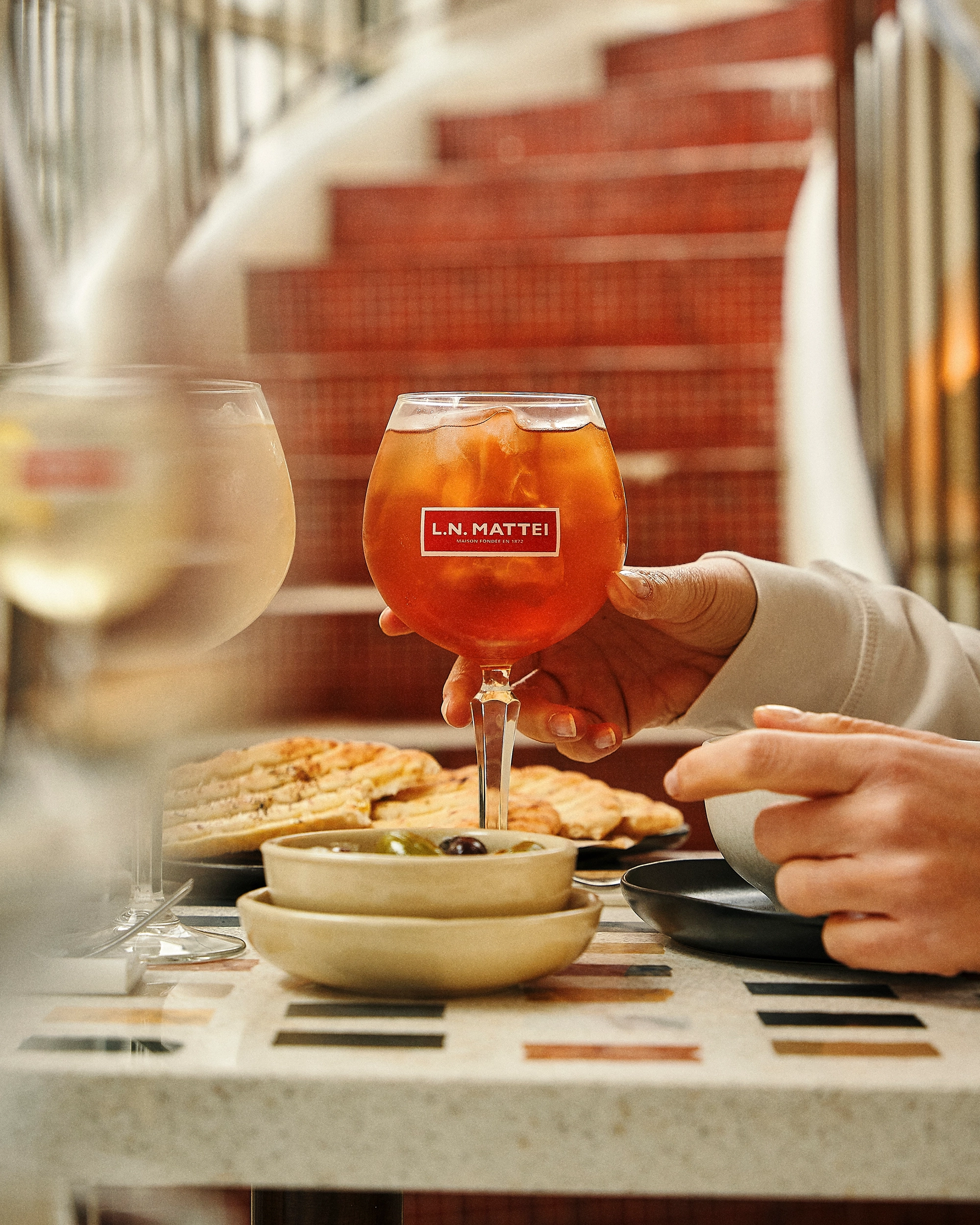 Person holding a glass of iced orange drink labeled L.N. MATTEI with plates of olives and flatbread on a patterned table near a red staircase.
