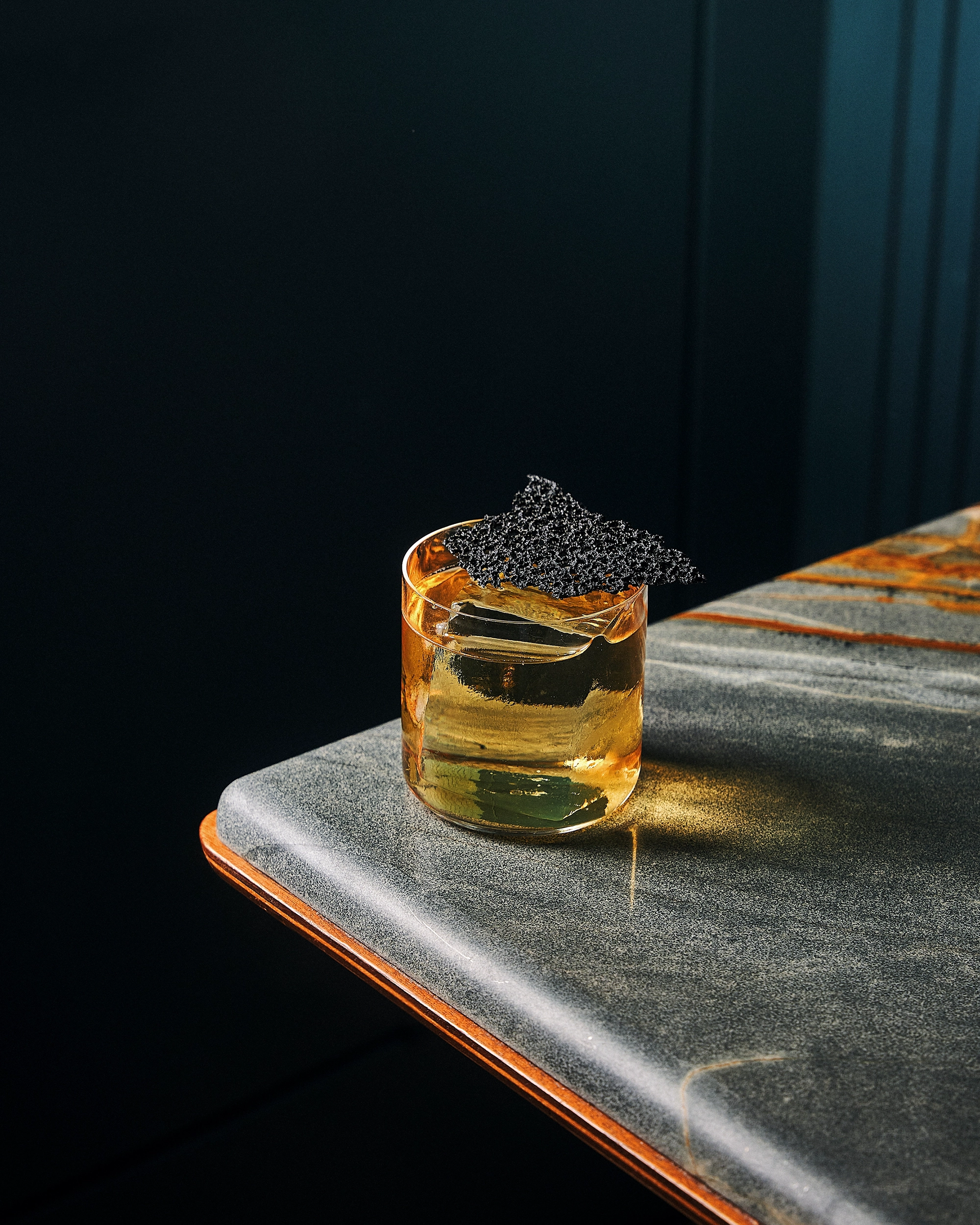 Glass of amber cocktail with a large ice cube and a black textured garnish on a gray marble table corner.