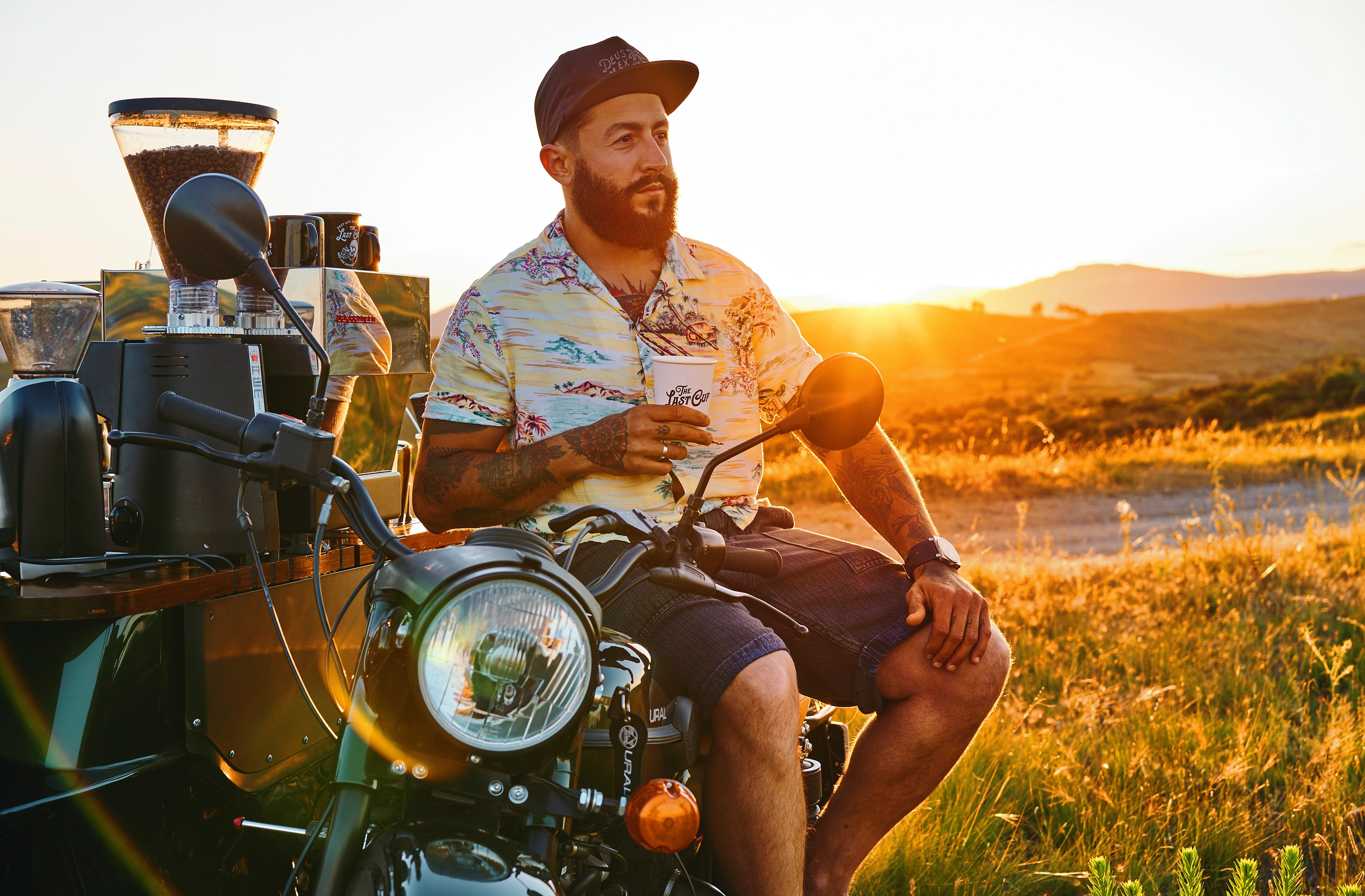 Bearded man in a floral shirt sitting on a motorcycle holding a coffee cup with a coffee machine behind him at sunset in a grassy field.