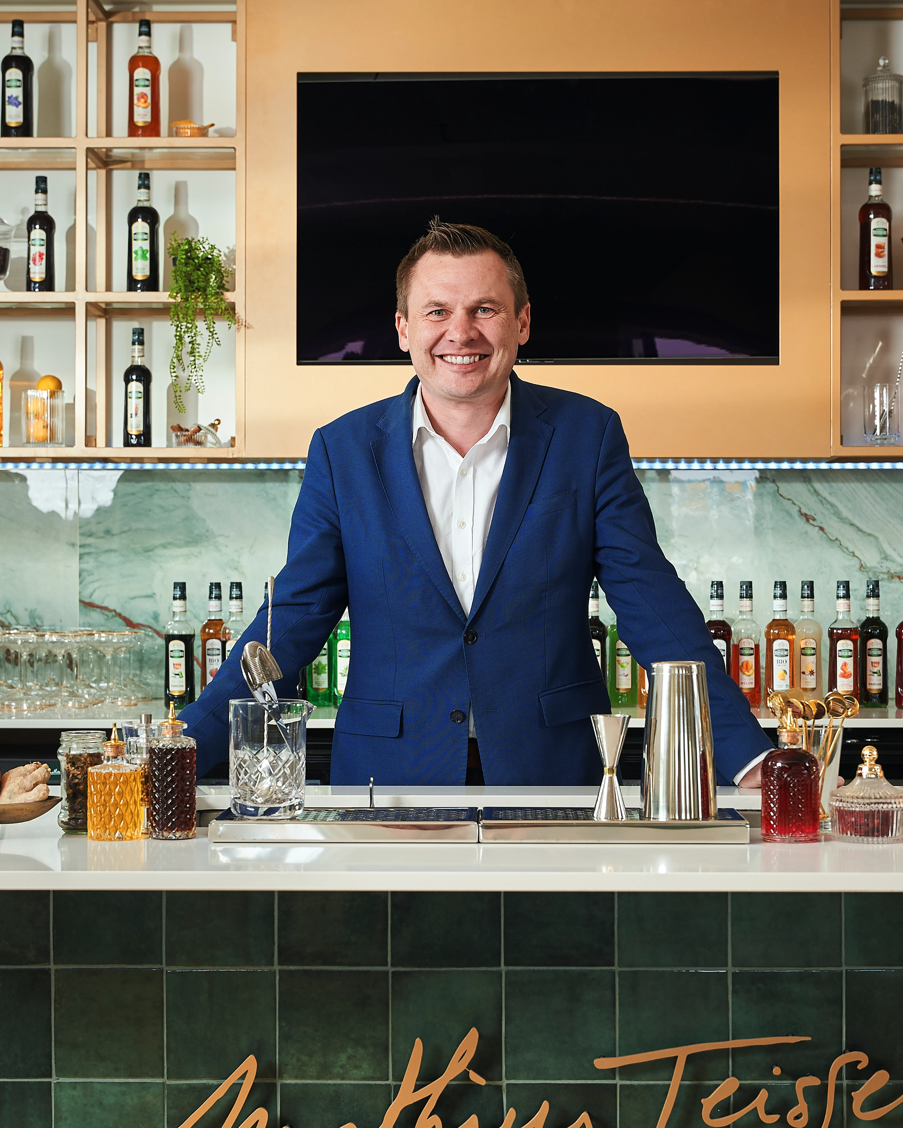 Smiling man in a blue suit standing behind a bar with bottles and cocktail tools, with a large black screen and shelf of liquor bottles in the background.