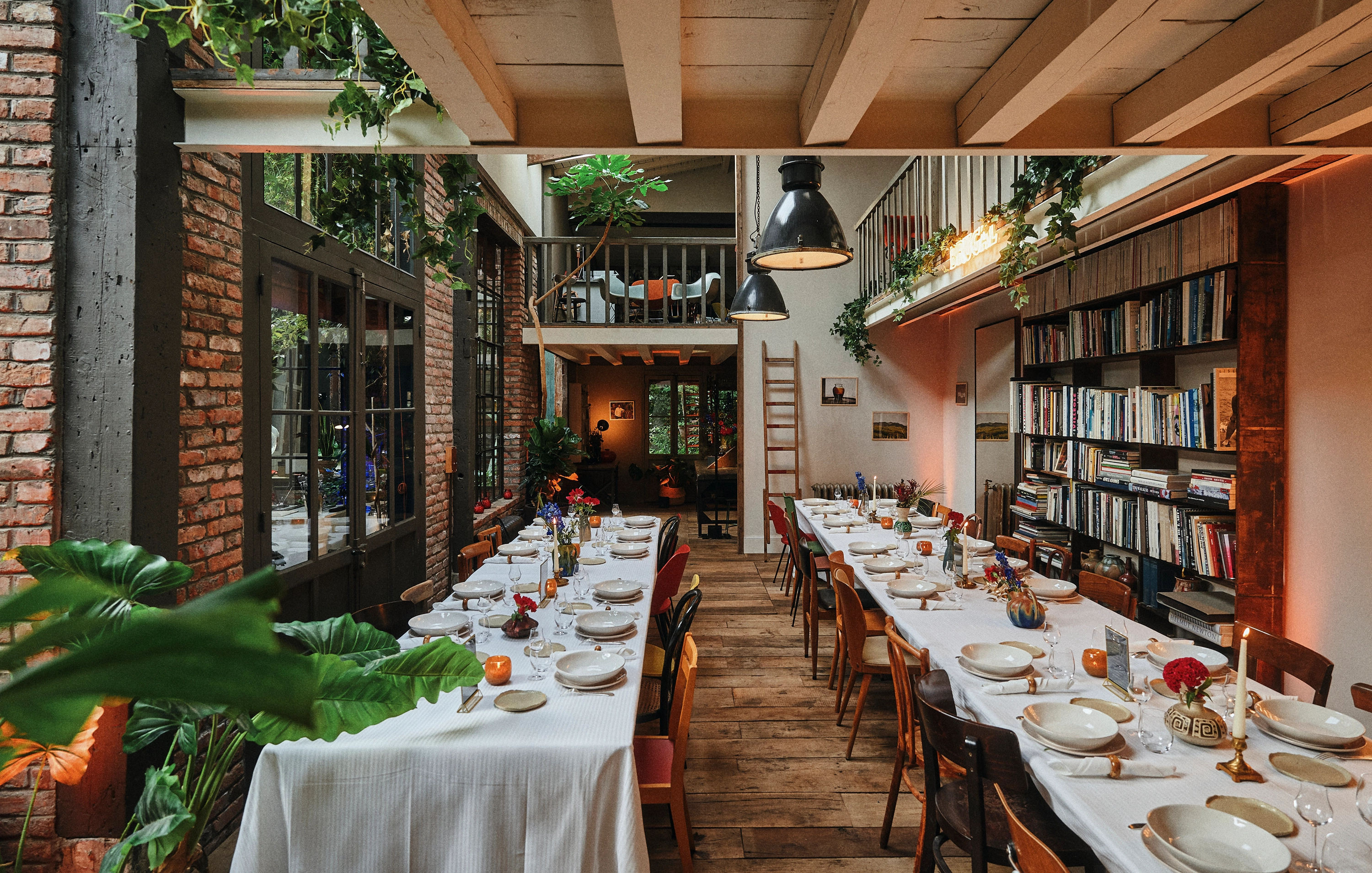 Long dining tables set with white tablecloths, plates, glasses, and candles in a rustic room with exposed brick walls, wooden floor, hanging plants, and a bookshelf.