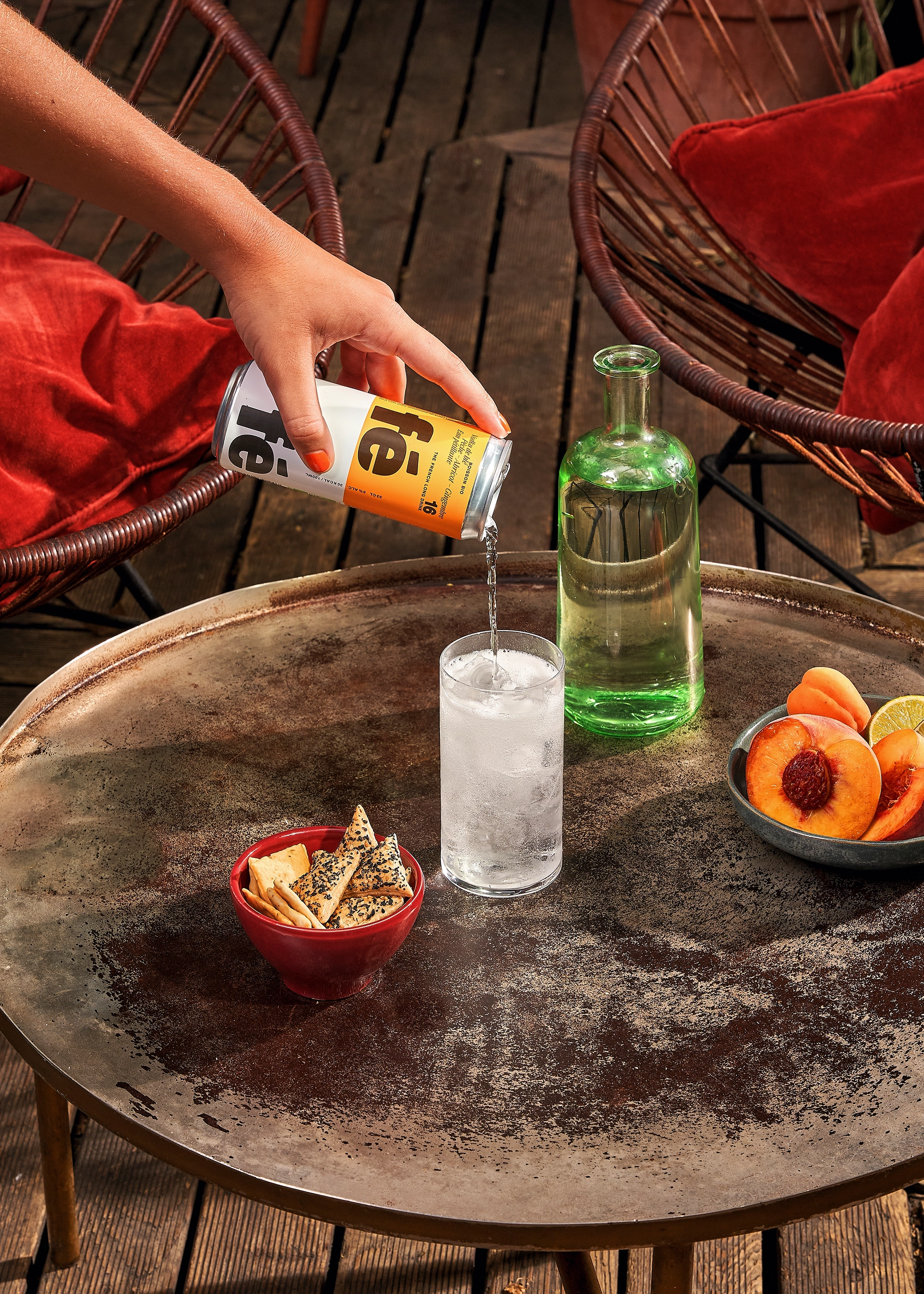 Hand pouring a fizzy drink from a can into a glass with ice on a rustic round table with bowls of crackers and sliced peaches.