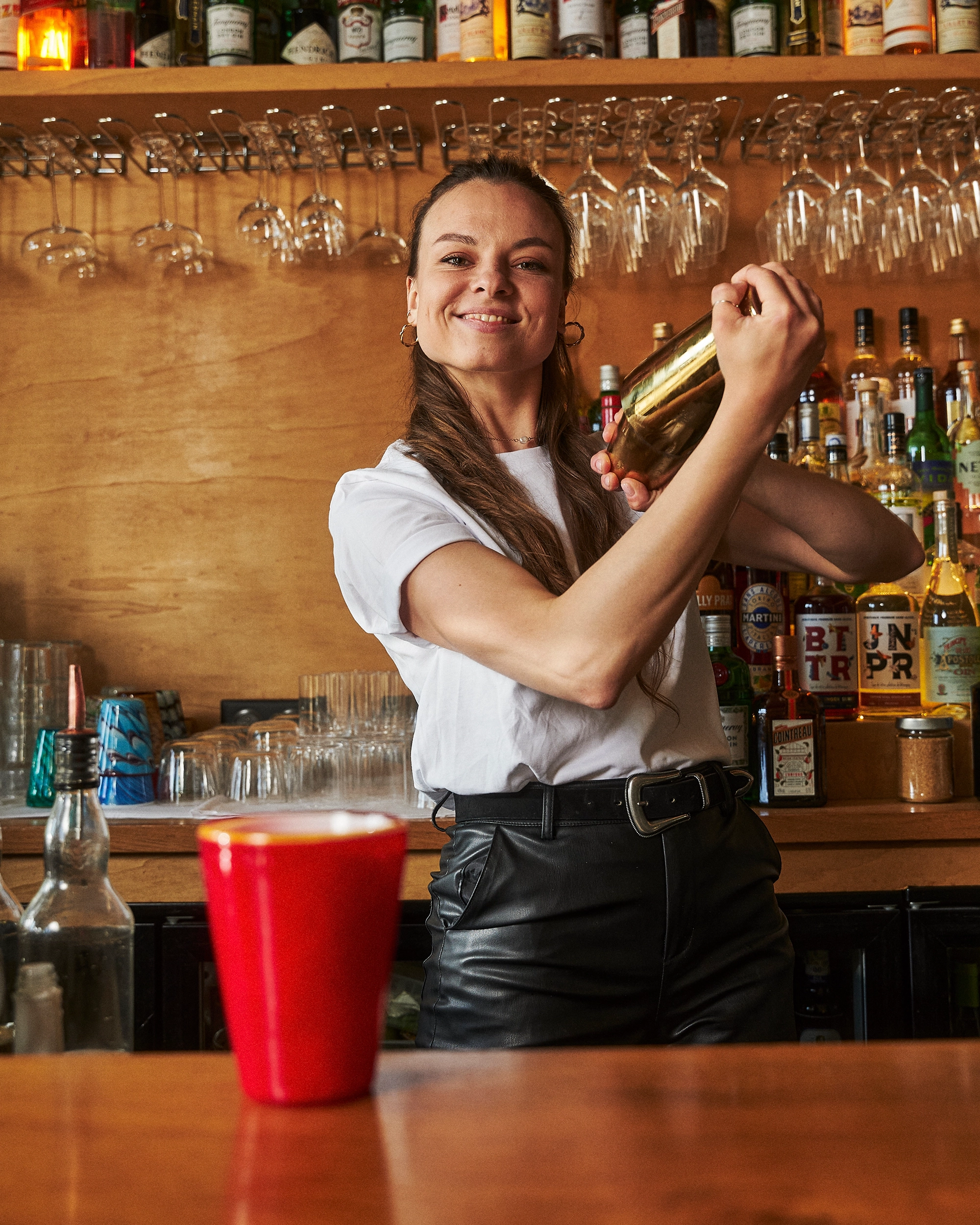 Smiling bartender shaking a cocktail shaker behind a bar with bottles and hanging glasses, red cup in the foreground.