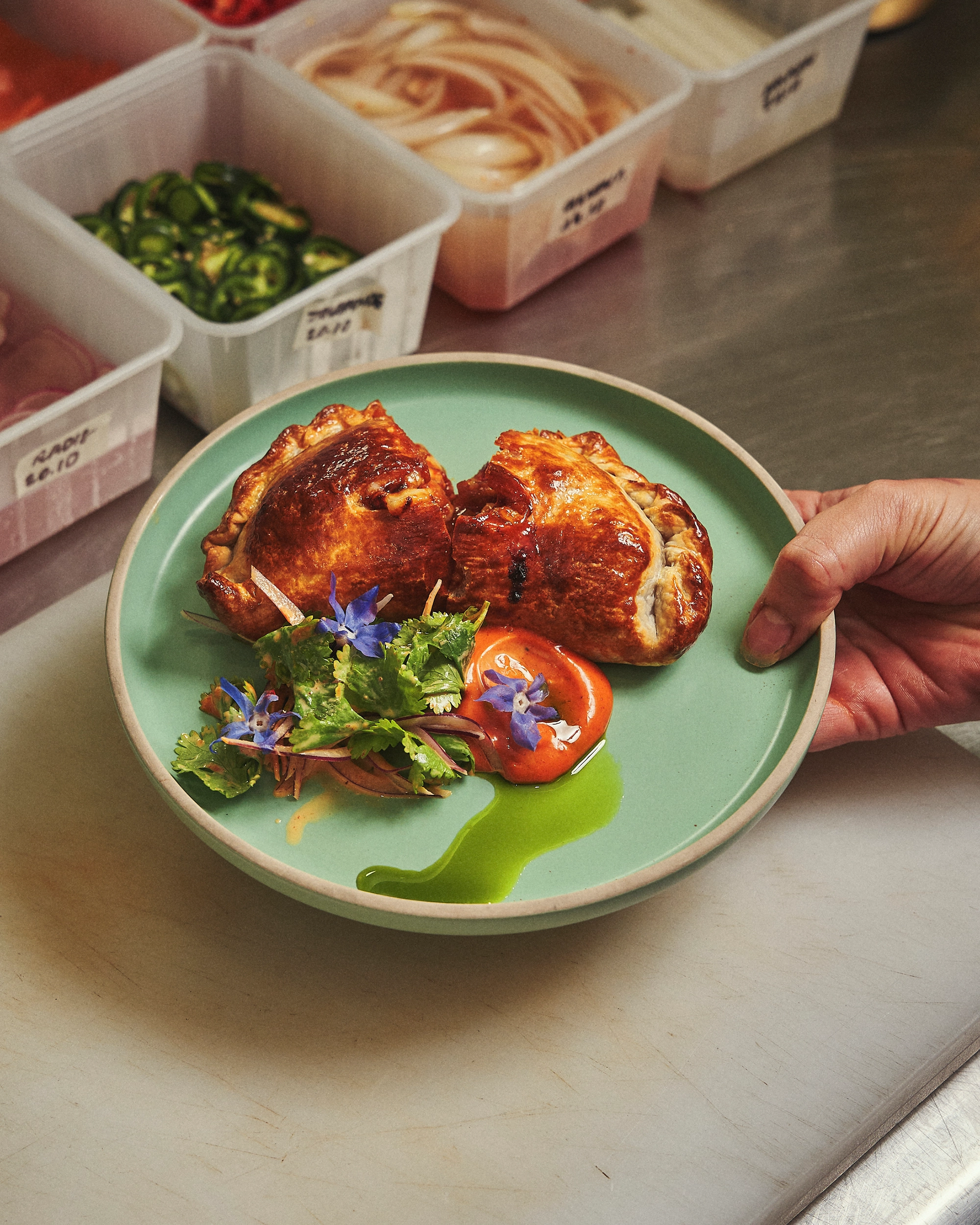 Hand holding a green plate with two golden-brown baked pastries, a dollop of red sauce garnished with blue flowers, green sauce, and a small herb salad.