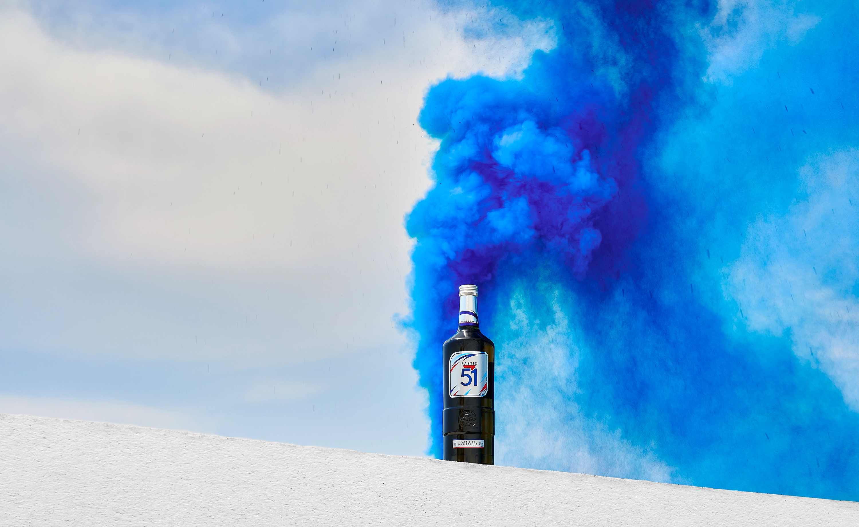 Bottle of Pastis 51 placed on a white surface with vibrant blue smoke swirling in the sky background.