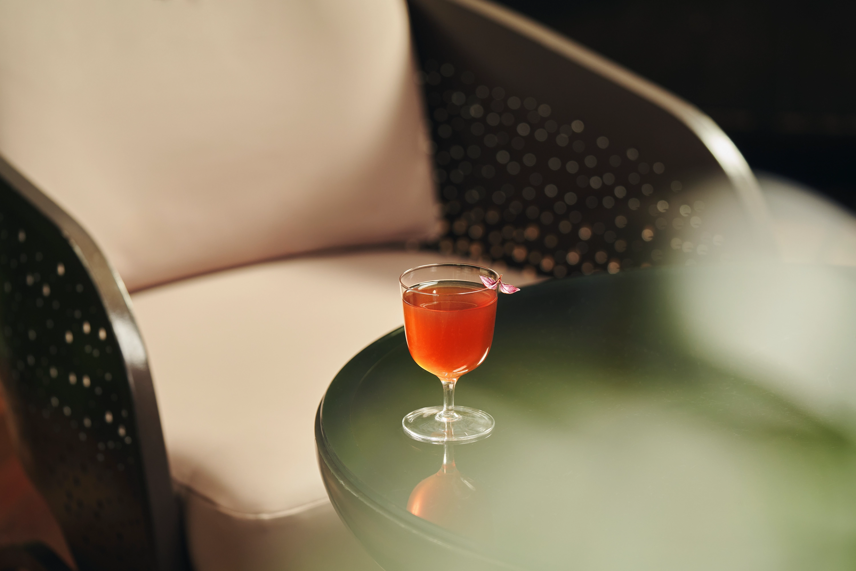 Orange cocktail garnished with a small pink flower on the rim, placed on a green table next to a light beige cushioned chair.