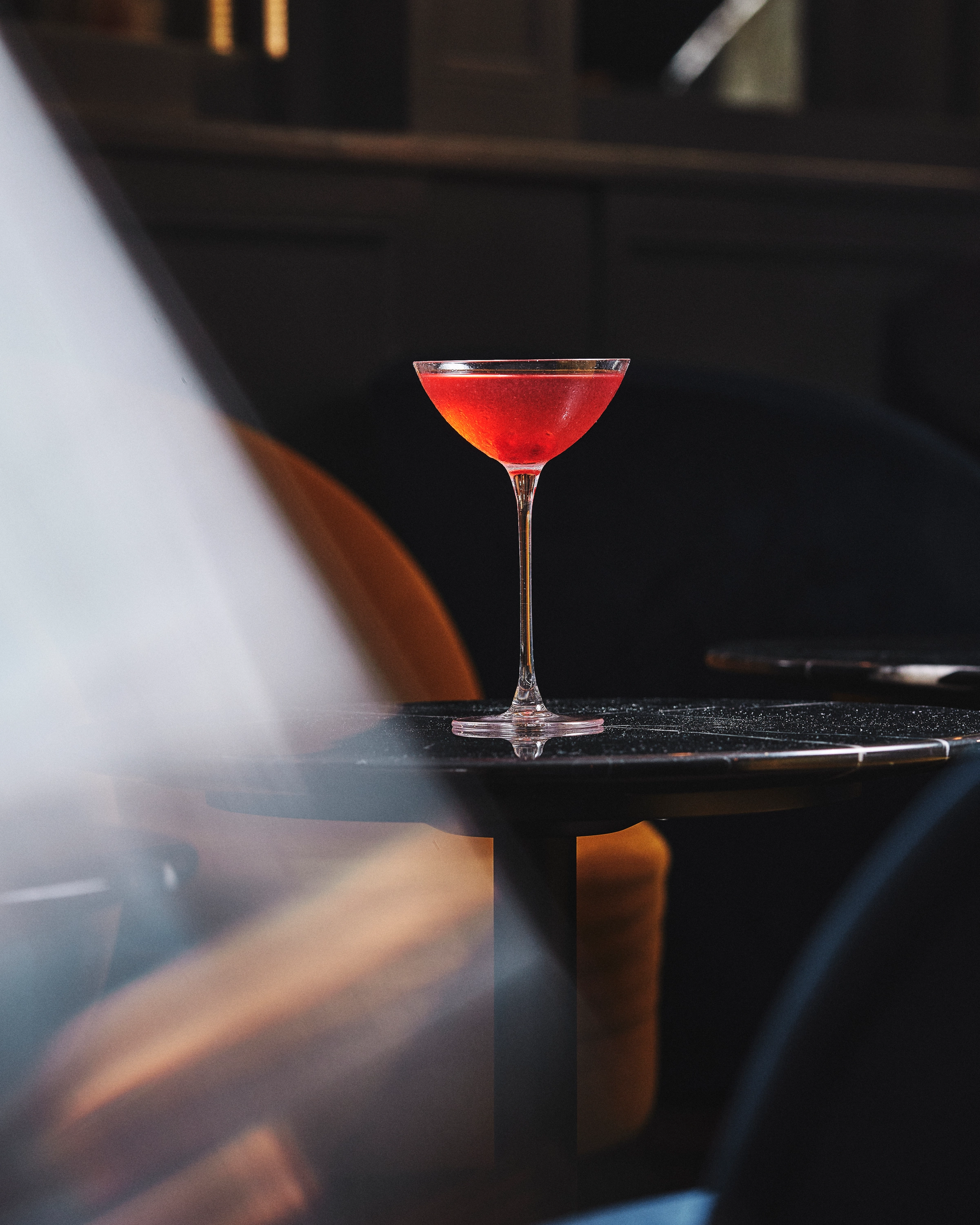 A red cocktail served in a coupe glass on a dark round table with blurred armchairs in the background.