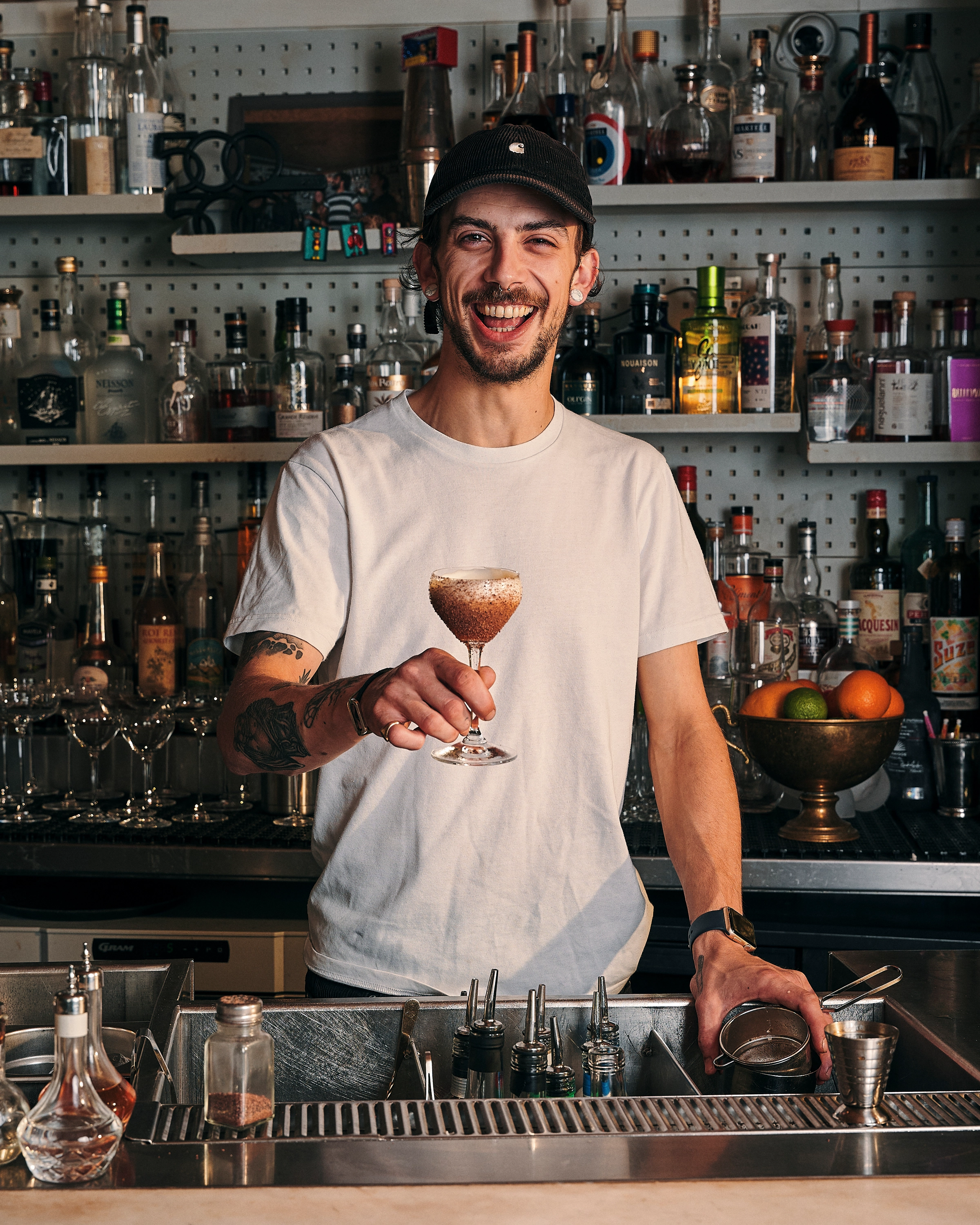 Smiling bartender in a white t-shirt and black cap holding a cocktail glass behind a bar with bottles and fruit in the background.