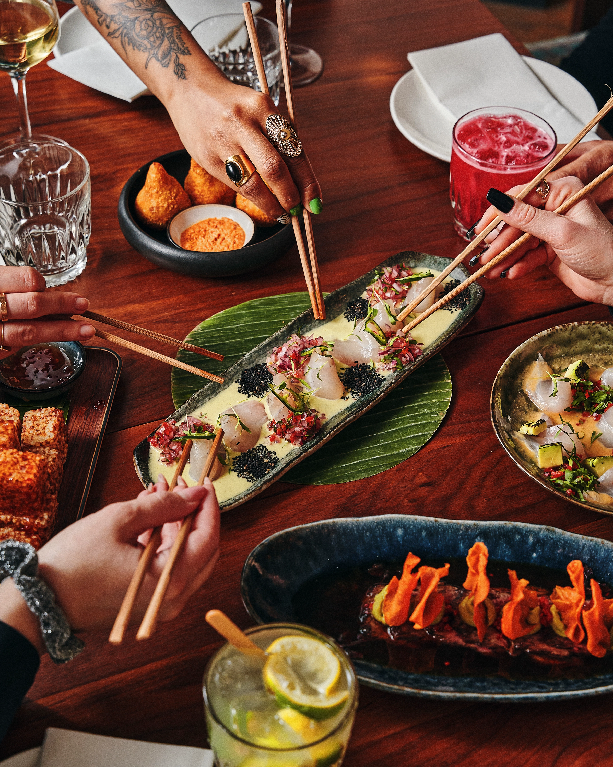 Four hands using chopsticks to pick pieces of sushi and sashimi from various plates on a wooden table with drinks and dipping sauce.