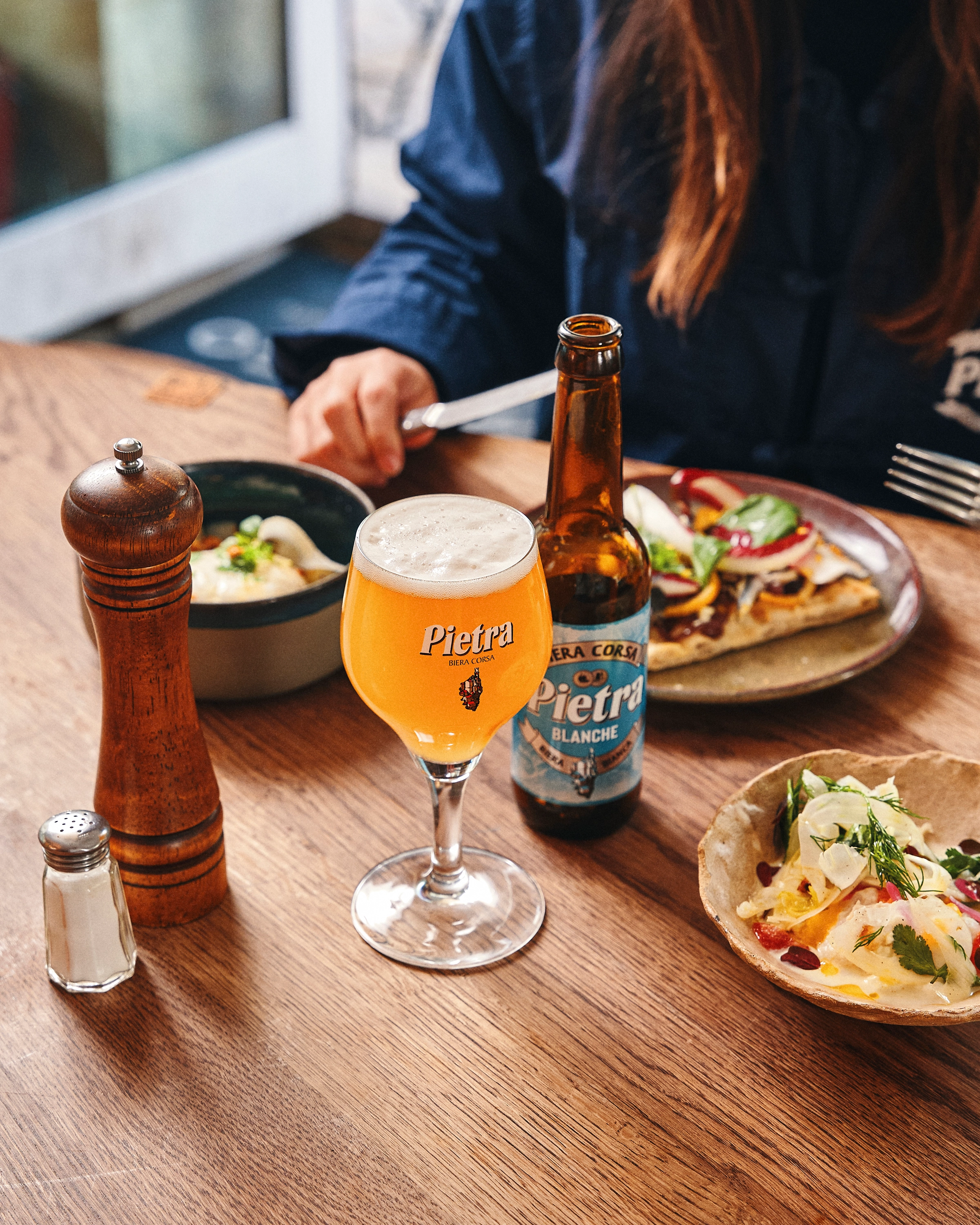 Glass of Pietra Blanche beer with bottle, salt and pepper shakers, and plates of food on a wooden table with a person in the background.