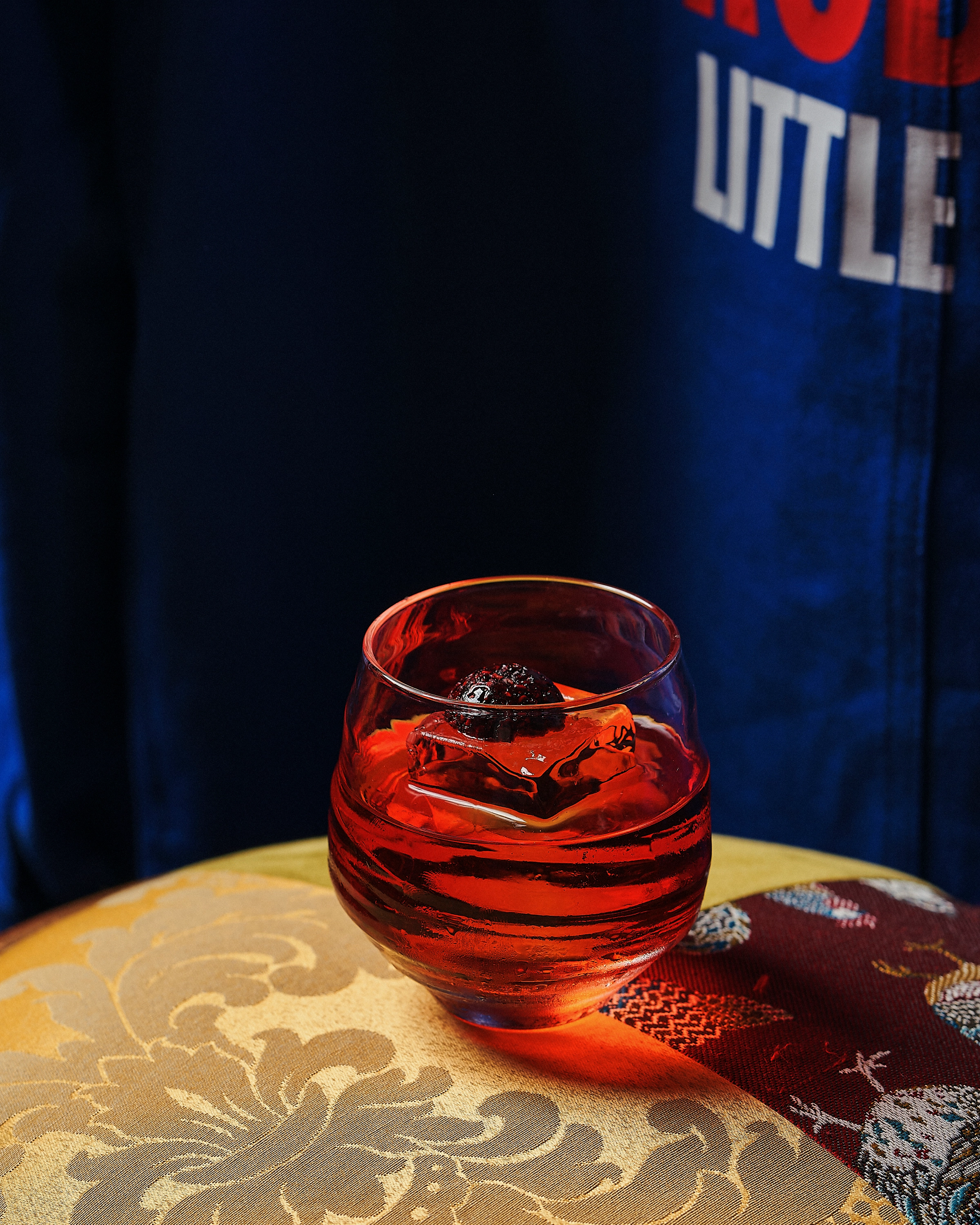 Red cocktail with a large ice cube and a dark garnish on top, placed on a patterned golden-yellow surface with a red cloth nearby.