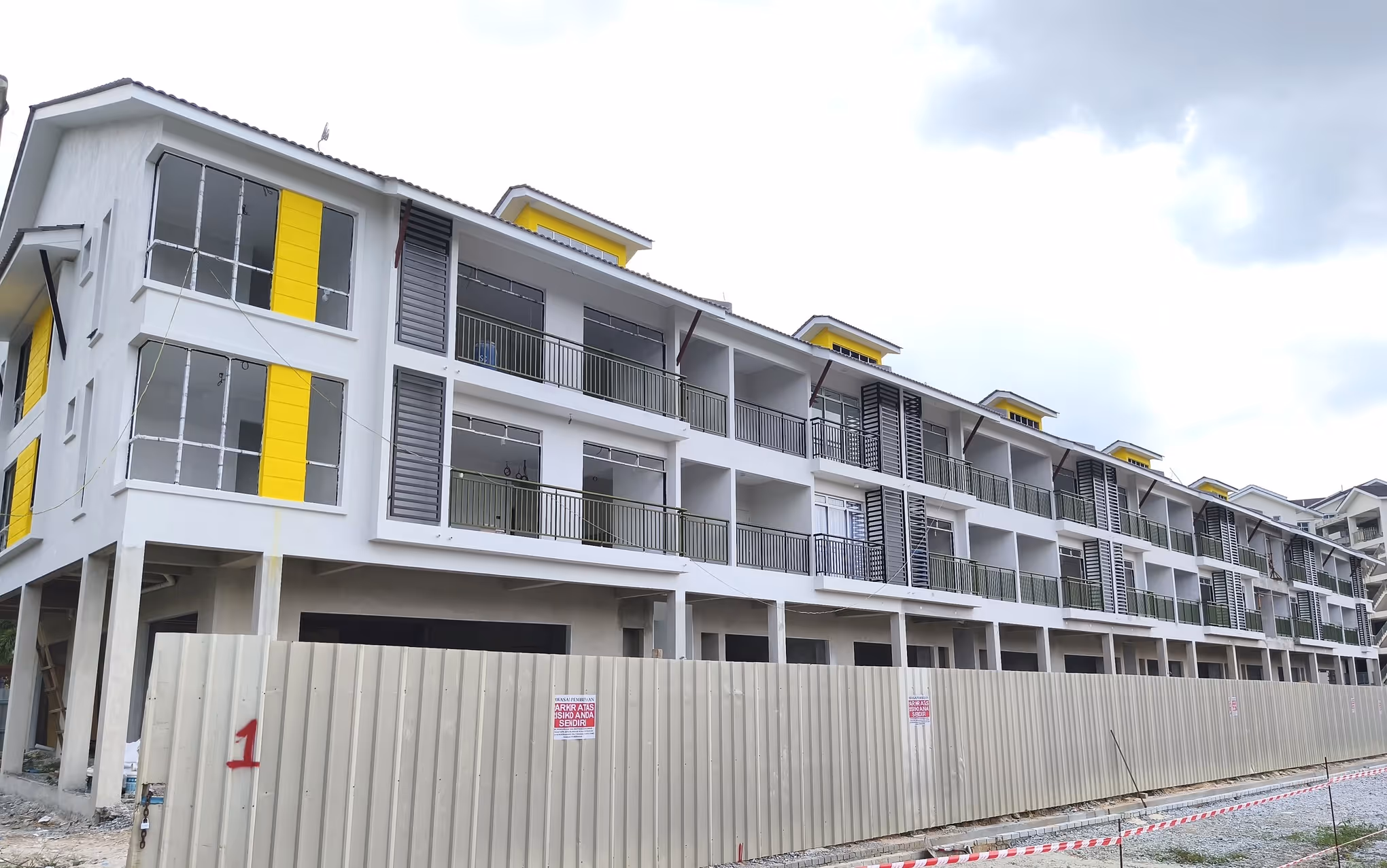 Three-story white building with yellow accents and balconies behind a construction fence on a cloudy day.