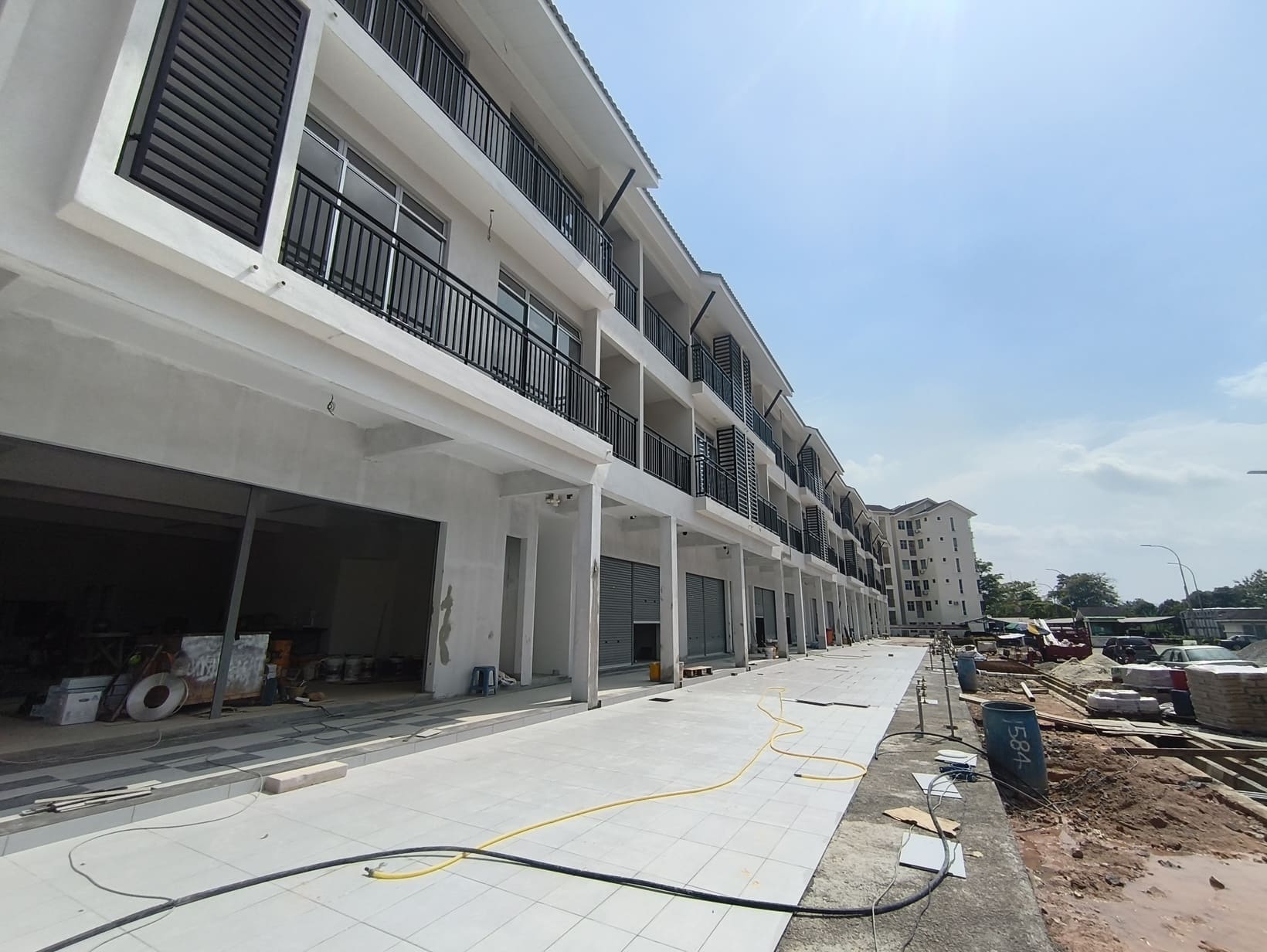 Three-story white building with yellow accents and balconies behind a construction fence on a cloudy day.