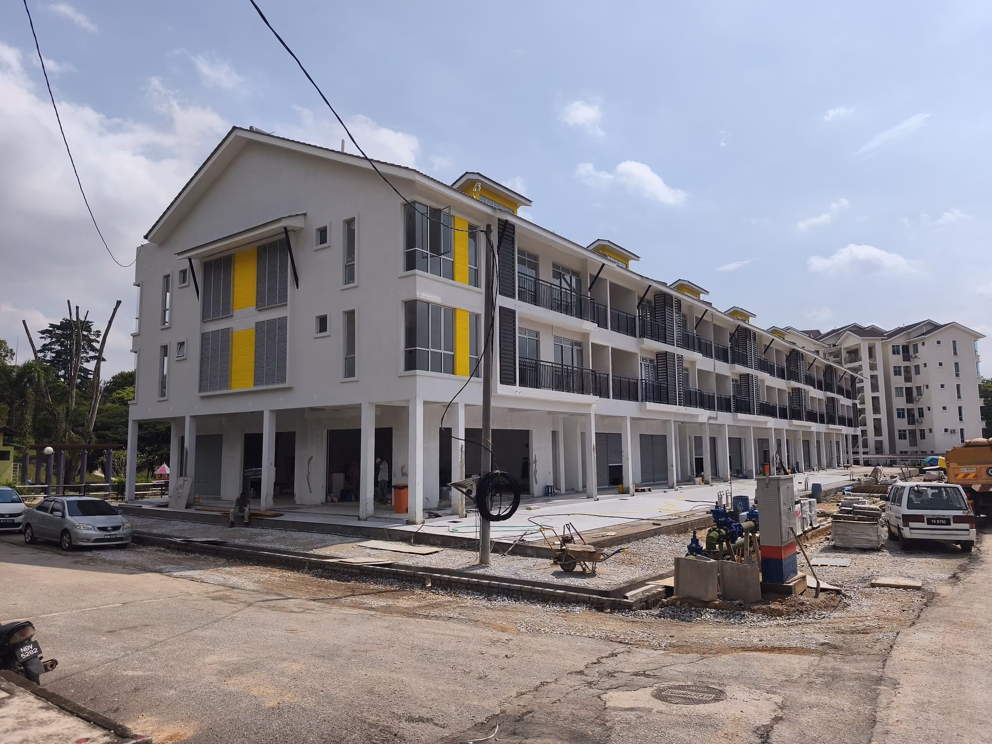 Three-story white building with yellow accents and balconies behind a construction fence on a cloudy day.