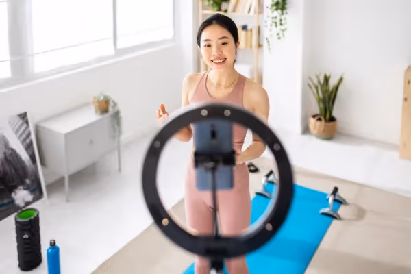 A cheerful young woman wearing activewear is standing in a bright, home exercise space, greeting an audience through a ring light that frames a recording device. The room is equipped with workout gear, including a yoga mat, dumbbells, and a water bottle, suggesting she is either about to start or has just finished a fitness session. Houseplants and minimalist furniture create a serene and tidy backdrop