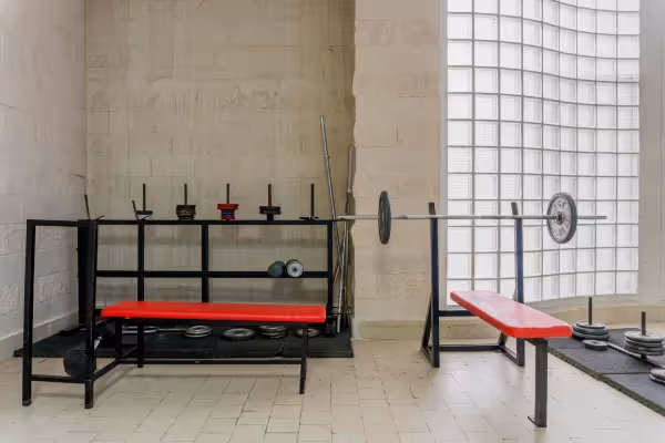A weightlifting area inside a gym. There is a squat rack with barbell supports and multiple weight plates on the rack and on the floor. A flat bench with a red surface is in the foreground, and another bench is positioned near the barbell, which is loaded with weights and ready for use. The space is well-lit, with natural light coming through a large grid window, and the walls are a neutral, light color, giving the room a clean and minimalistic appearance. The gym appears to be empty, emphasizing the equipment and the readiness of the space for a workout session.