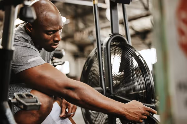 A man in a gym setting, focused intently on his workout. He is seated, wearing a gray t-shirt, which highlights his muscular build. The man is holding onto a piece of gym equipment, possibly taking a rest between sets or focusing before beginning an exercise. A large industrial fan is visible in the background, indicating a well-ventilated space, often necessary in a fitness environment to help keep the area cool during intense physical activity. The image captures a moment of concentration and physical exertion, common in fitness and weight training activities.