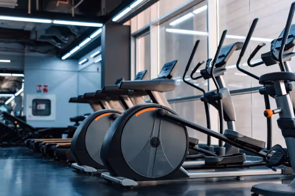 A row of elliptical trainers in a gym. These machines are designed for cardiovascular exercise and feature large, round bases with foot pedals and upright handles, which users push and pull in a gliding motion. The ellipticals are positioned next to a window, allowing for natural light to enter the space. Above the machines, there's a series of fluorescent lights, contributing to the well-lit environment. The walls are a neutral grey, and the floor seems to be made of a durable material suitable for heavy gym equipment. This setup is typical for a modern fitness center, where such machines are lined up to face outward, possibly for users to enjoy the view or watch monitors during their workout. The gym appears clean and well-maintained, ready for users to engage in their fitness routines