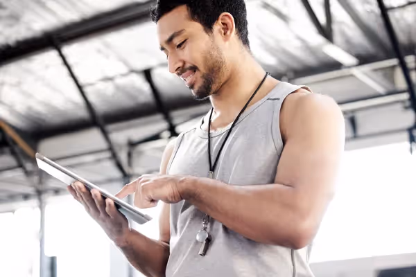 A man in a sleeveless gray top, likely in a gym setting, focused on a tablet he is holding. He has a lanyard around his neck, which could suggest he is a gym instructor or personal trainer tracking workout routines or schedules. The background is slightly overexposed, with high ceilings and industrial features, consistent with modern gym aesthetics. His engagement with the tablet and his attire indicate he might be planning a workout, reviewing client progress, or possibly using a fitness app to aid in training