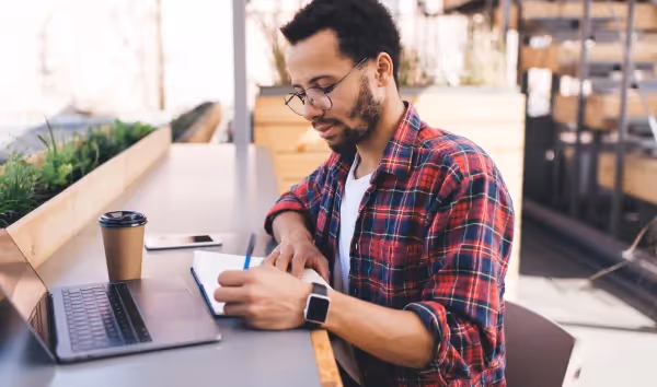 A person with glasses and a plaid shirt is focused on writing in a notebook, sitting at an outdoor table with a laptop and a cup of coffee next to them. They are wearing a smartwatch, and there appears to be a blurred urban background suggesting a casual work or study environment