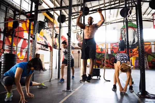 A group of people are engaging in various exercises at a CrossFit gym. The foreground shows a person in a blue top doing a push-up, while another in a polka dot tank top is preparing to lift a barbell. In the background, a shirtless individual with athletic build is lifting weights overhead, and another is bending to touch their toes. The gym is decorated with vibrant graffiti art on the walls, adding an urban, energetic vibe to the scene