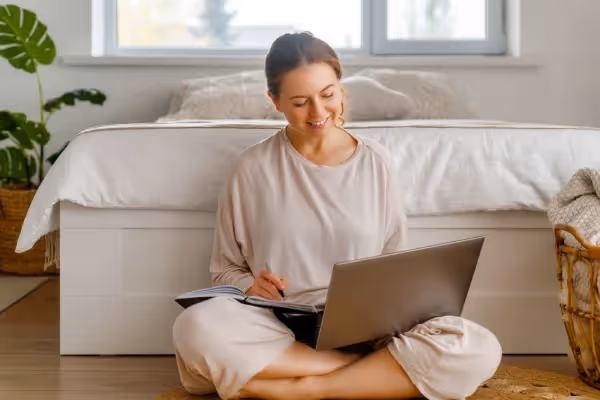 A woman sitting cross-legged on the floor, smiling while working on a laptop that is resting on her lap. She is casually dressed in a comfortable, pale outfit, and holding what appears to be a pen or stylus, possibly interacting with the laptop or taking notes. The setting is a bright, cozy room with a bed and plants in the background, suggesting a relaxed home environment