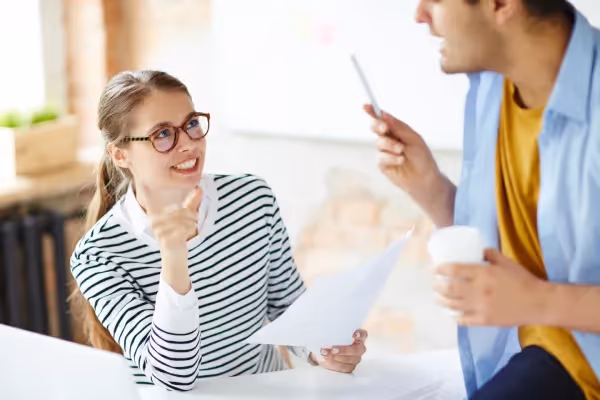 A woman in a striped shirt and glasses is engaged in a lively conversation with a man whose back is to the camera. She appears animated and possibly explaining a point or idea, while the man is holding a smartphone and a coffee cup, suggesting a casual meeting or discussion. The setting looks bright with a blurred background, indicating an informal office or co-working spac