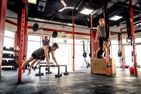An indoor gym where three people are engaged in different types of exercises. On the left, there's a person doing an overhead lift with a barbell, in the center, another individual is using parallel bars likely for push-ups or dips, and on the right, someone is standing atop a plyometric box, which might be used for box jumps or step-ups. The gym has a variety of equipment, suggesting a focus on functional fitness or CrossFit-style workouts. The environment is well-lit and appears to be designed for high-intensity training with ample space for movement and a variety of exercises