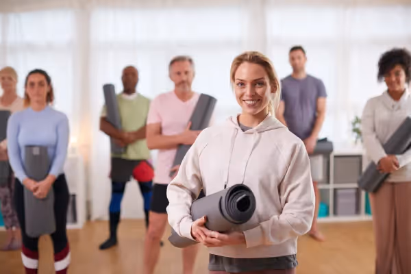 A group of adults in a yoga or fitness class setting. In the foreground, a smiling woman with her hair tied back is holding a rolled-up yoga mat, wearing a casual, light-colored hoodie. Behind her, the rest of the class members are standing, each holding their own yoga mats, ready for the session. They are wearing various exercise clothes, indicating a diverse group of participants. The group's attention seems to be towards the front, possibly listening to an instructor or preparing to start the class. The room is bright and airy, creating a welcoming and calm atmosphere suitable for a yoga or fitness class