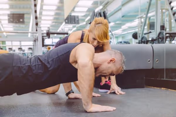 Two people in a gym setting. A man is in the foreground doing a push-up, and a woman is closely observing or assisting him. The woman appears to be in a coaching or spotter position, possibly providing feedback or encouragement. They are both focused on the exercise at hand. The environment is a well-equipped gym with various machines and mirrors in the background, indicating a place dedicated to fitness and training. The man is wearing a black outfit and a watch, which could be a fitness tracker, while the woman sports a purple top, suggesting they are dressed appropriately for a workout session. The overall impression is one of a personal training session or a focused workout with attention to form and technique.