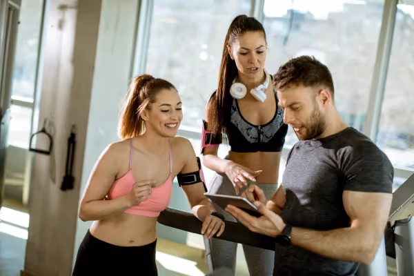 Gym setting where two women and one man are interacting with each other. The man is holding what looks to be a tablet and is pointing at the screen, possibly showing some information or data. One woman, wearing a pink sports top and black leggings, seems to be in motion as if she is running or walking on a treadmill and looking at the tablet. The other woman, dressed in a black and gray sports bra and leggings with headphones around her neck, is standing and looking attentively at the tablet as well. The environment suggests a focus on fitness and possibly technology integration into their workout, as evidenced by the use of a tablet and the wearable device on the woman in pink. The lighting is bright, and the mood seems positive and collaborative.