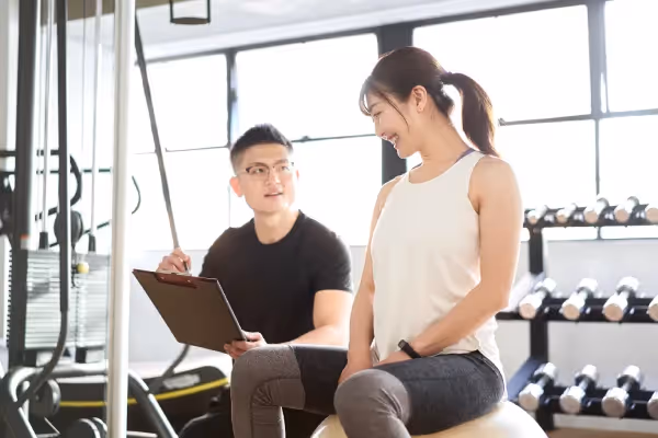 A personal training session in a gym, featuring a male trainer with a clipboard attentively looking at a female client sitting on an exercise ball, both are smiling, suggesting a positive interaction, with gym equipment and weights in the background