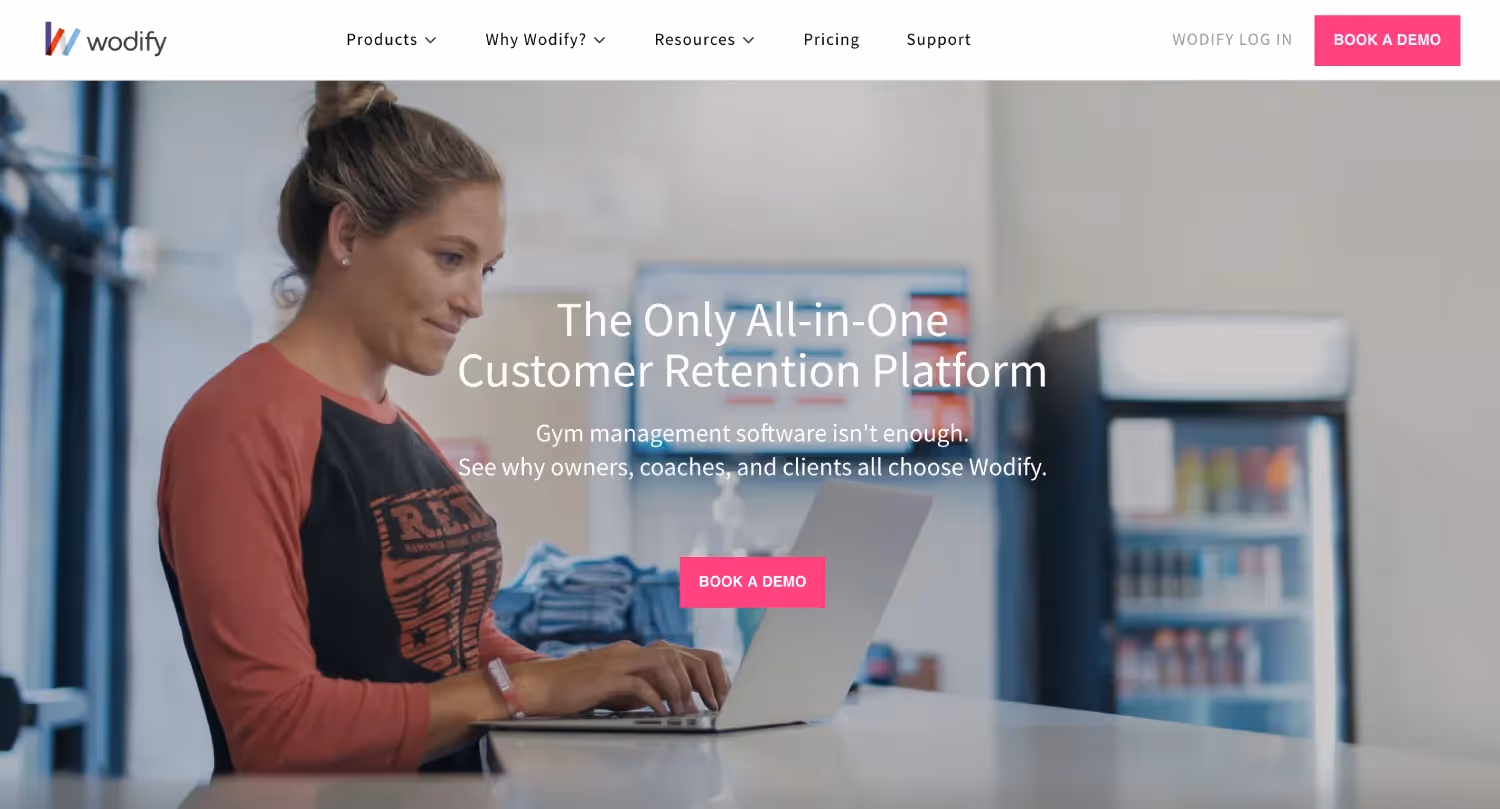 Screenshot of Wodify's homepage featuring a woman working on a laptop in a gym setting with text promoting it as the only all-in-one customer retention platform