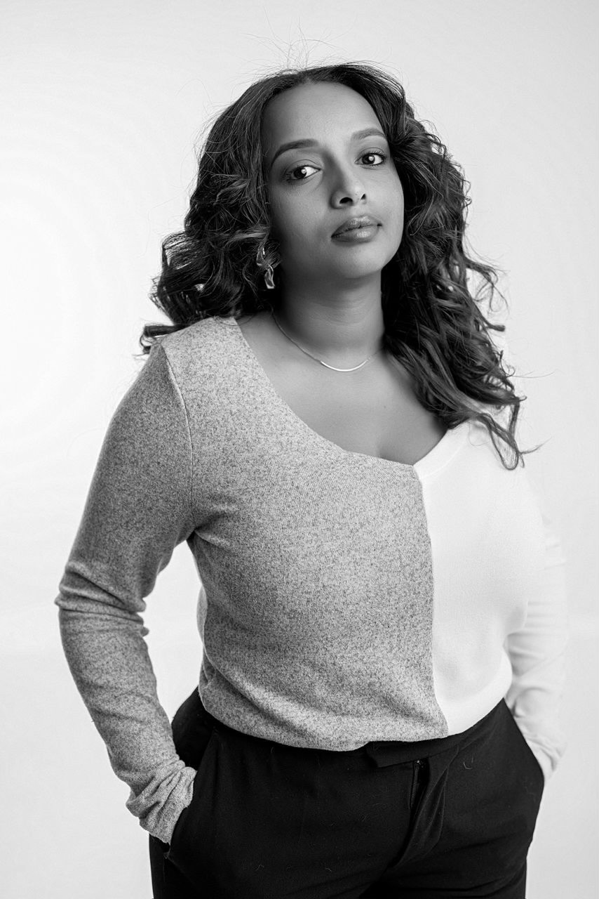 Portrait of a woman with curly hair wearing a two-tone sweater and black pants, hands in pockets, looking confidently at the camera.