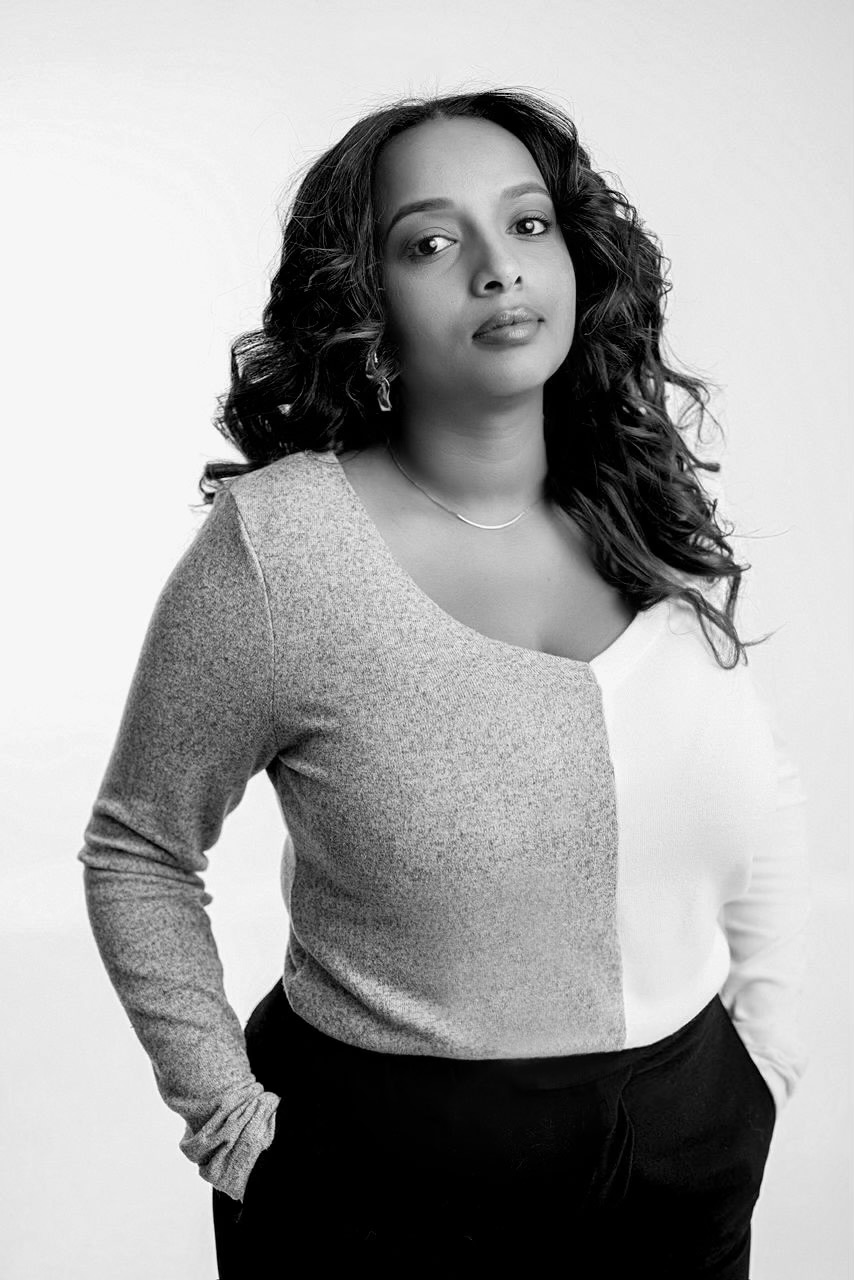 Black and white portrait of a woman with curly hair wearing a two-tone sweater and black pants, with hands in pockets.