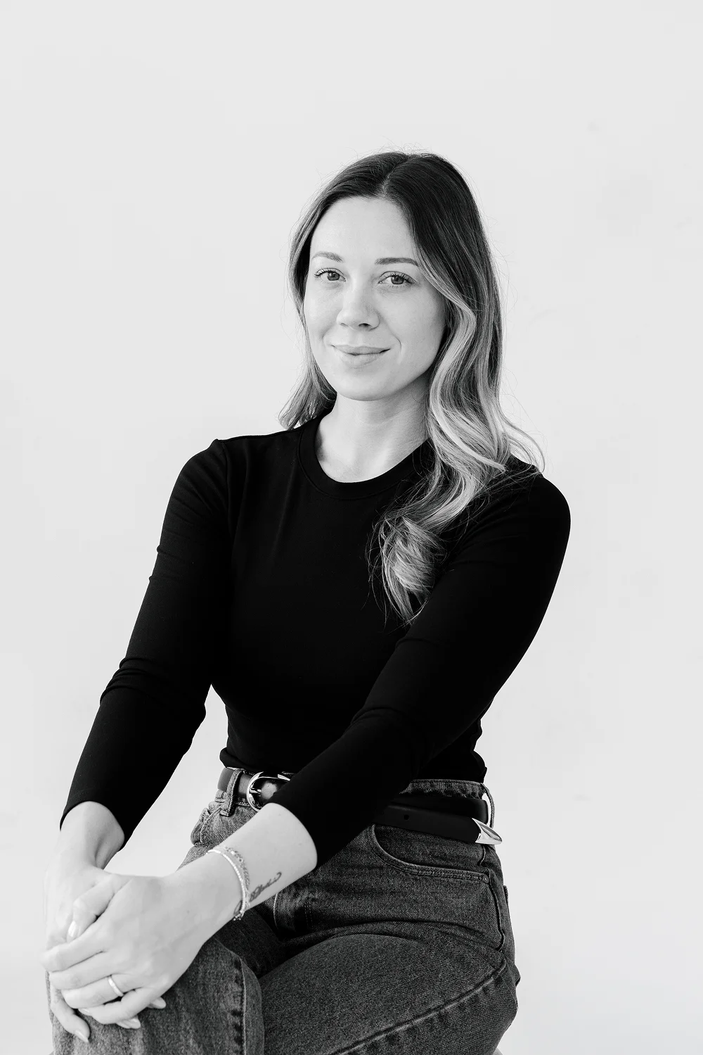 Smiling woman with long wavy hair wearing a black long-sleeve shirt and jeans, sitting with hands clasped.