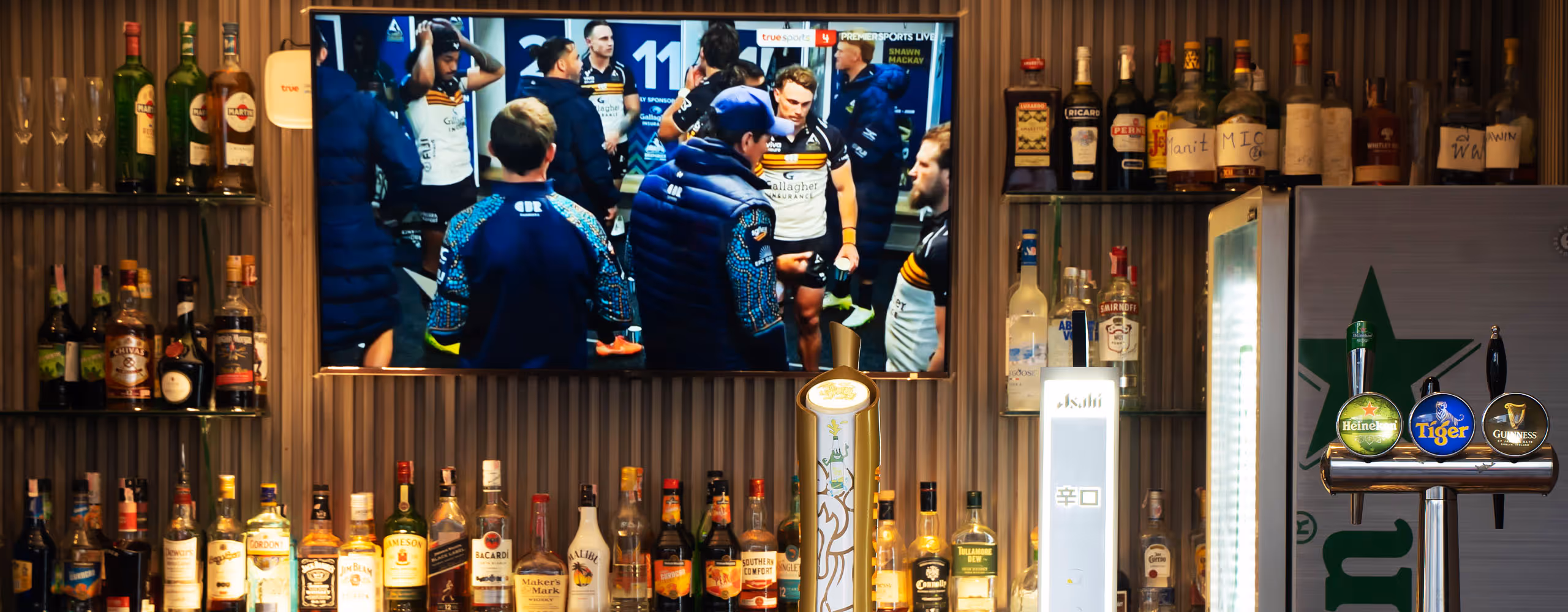 Bar with shelves of liquor bottles, four beer taps, and a TV showing a rugby team huddle.