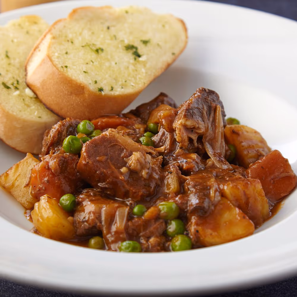 Beef stew with peas, carrots, and potatoes served in a white bowl with two slices of garlic bread.