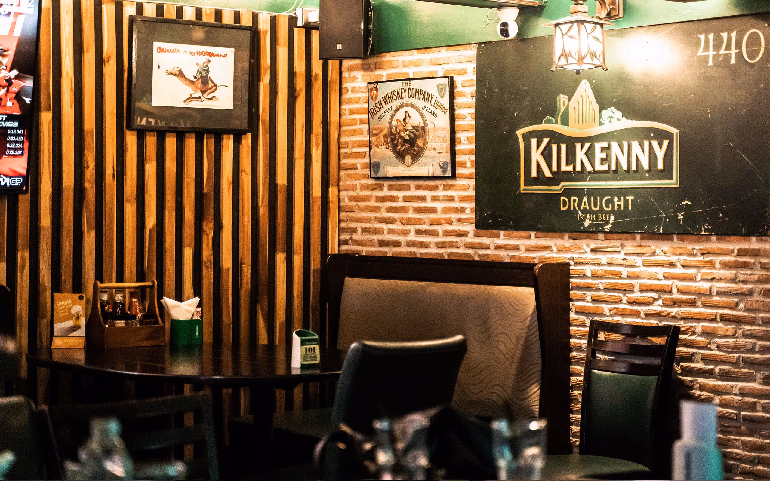 Cozy pub corner with wooden walls, framed vintage whiskey and beer signs, tables, chairs, and booth seating.