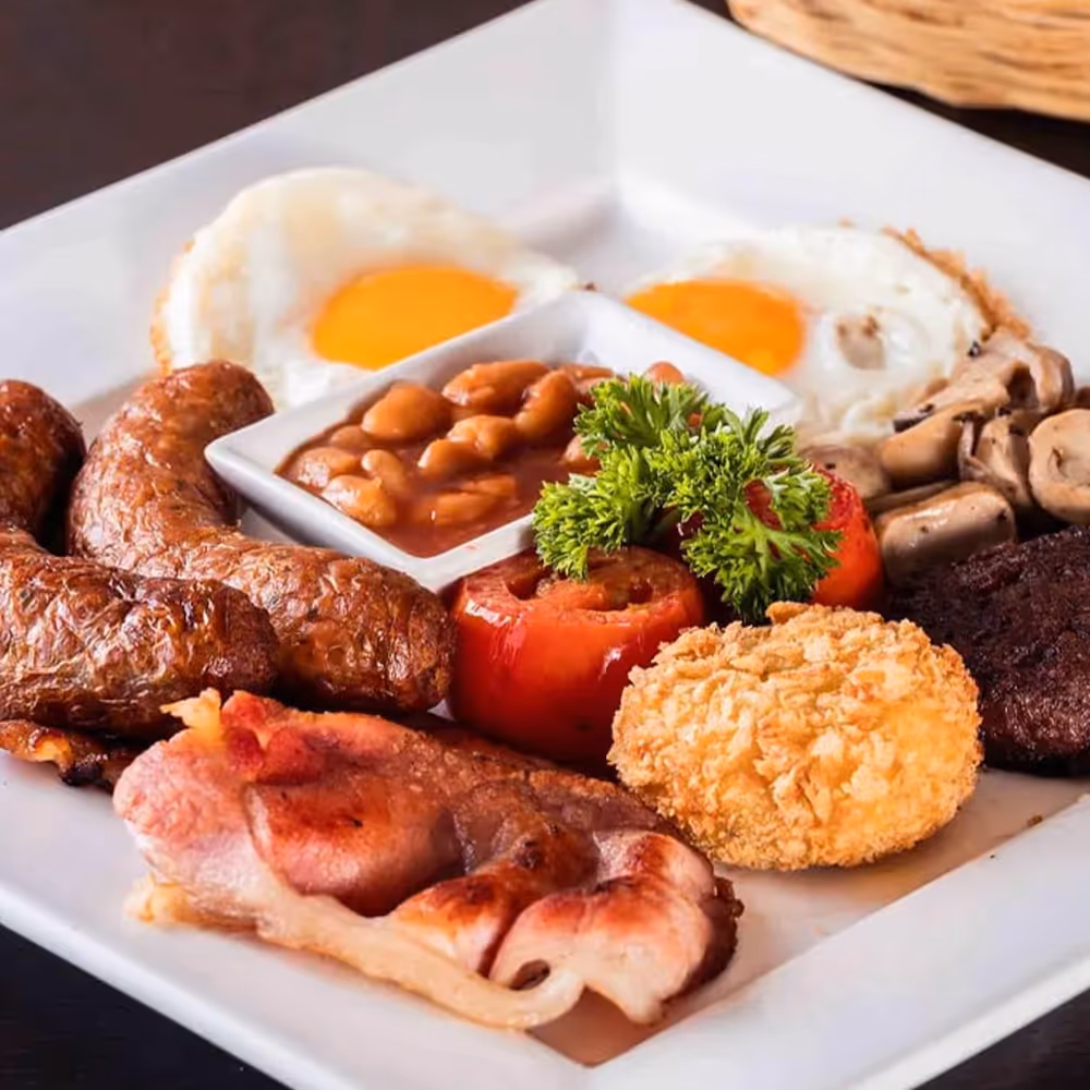 Full breakfast plate with fried eggs, sausages, baked beans, grilled tomato, bacon, mushrooms, and a fried item.