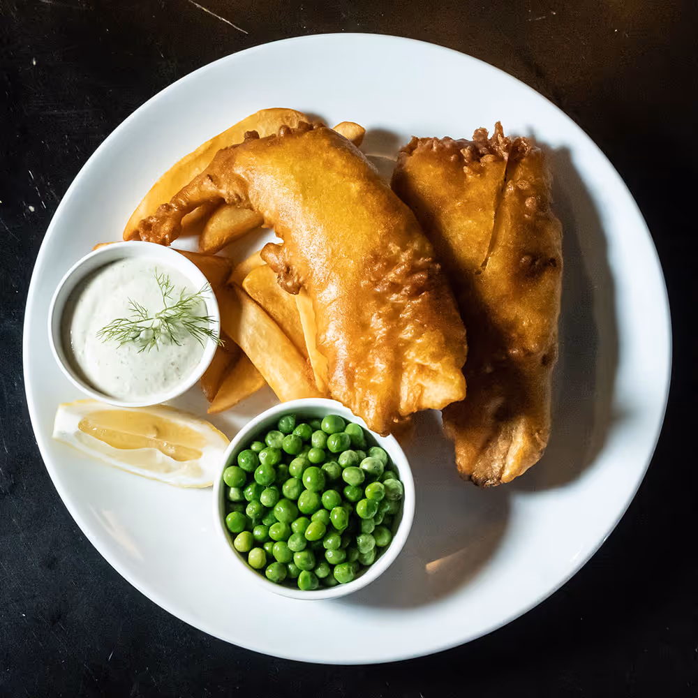 Plate of fish and chips with tartar sauce, lemon wedge, and a bowl of green peas.