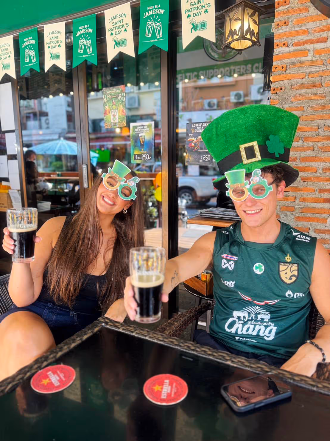 Two people wearing St. Patrick's glasses and hat, holding pints of dark beer at a pub.