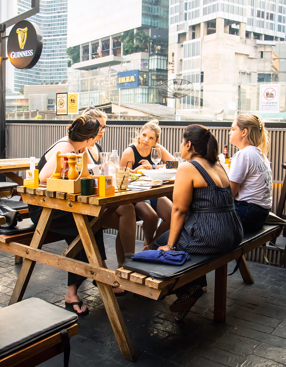 Five women sitting around a wooden picnic-style table at an outdoor urban restaurant.