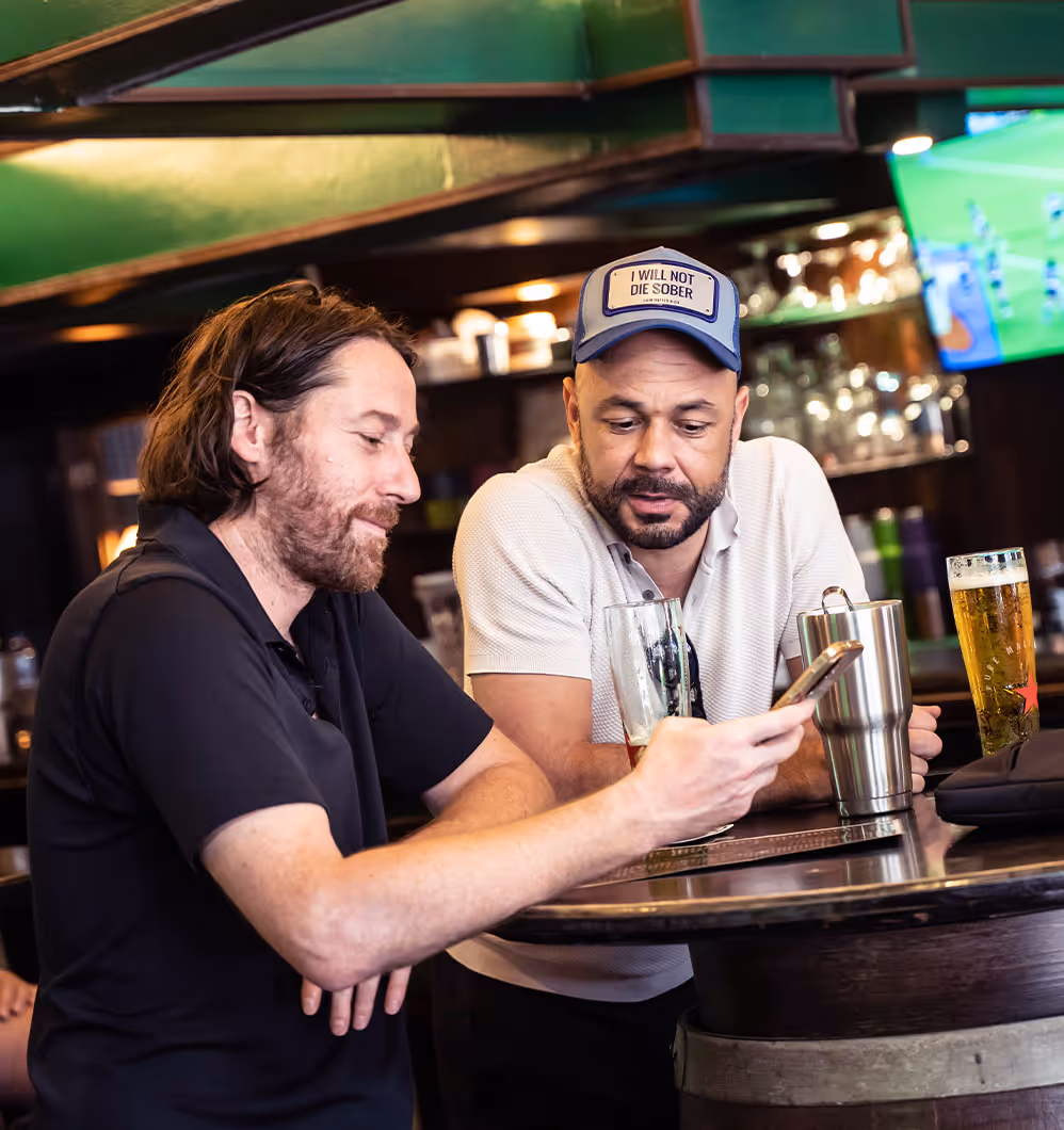 Two men sit at a pub table with drinks, one showing the other something on his phone.