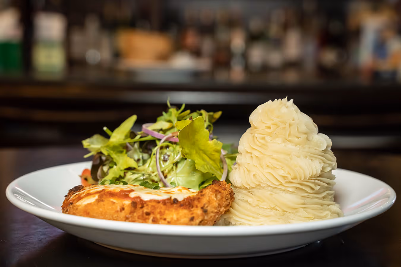 Plate with crispy chicken parmigiana, mashed potatoes shaped in a swirl, and mixed green salad.